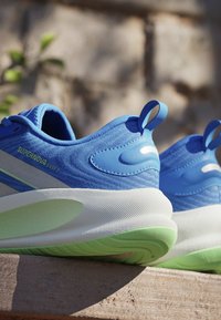 Paire de baskets athlétiques bleues et vertes avec des semelles blanches épaisses, avec le texte "SUPERNOVA EASE 2" sur le côté, placées sur une surface en bois.