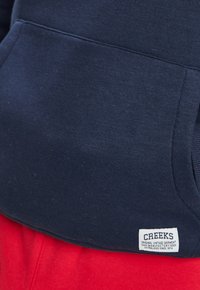 Felpa blu navy con tasca frontale, realizzata in un morbido cotone misto, caratterizzata da un'etichetta bianca con il testo "CREEKS" su uno sfondo rosso.