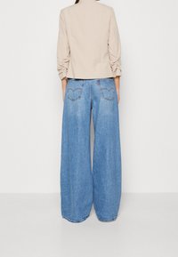 Chaqueta beige entallada con mangas fruncidas, combinada con vaqueros de mezclilla azul de pierna ancha con bolsillos traseros y lavado descolorido.