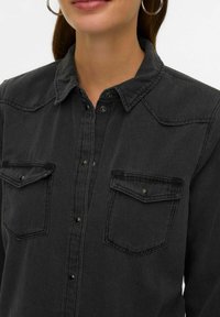 Czarna koszula jeansowa z zapięciem na guziki, zaostrzonym kołnierzem, dwoma kieszeniami na piersi i srebrnymi ozdobami. Tkanina o fakturze, luźny krój.