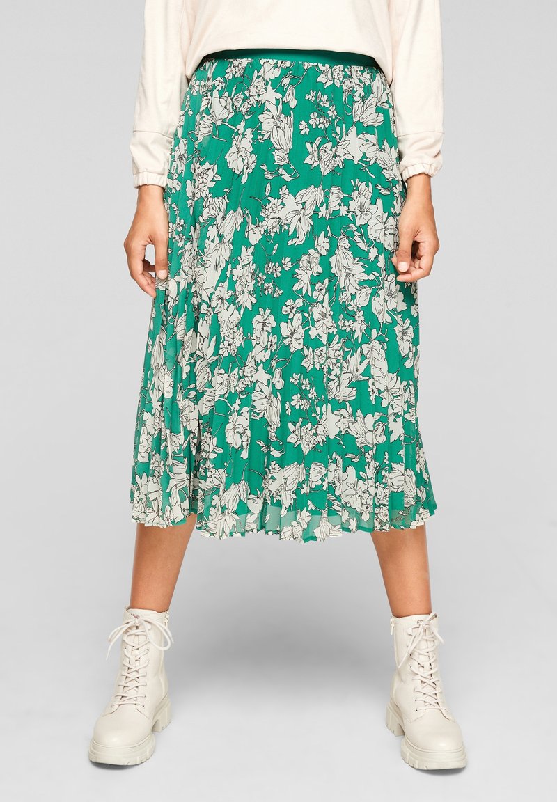 Femme portant une jupe midi plissée verte à fleurs avec des bottines blanches à lacets, debout devant un fond clair uni.