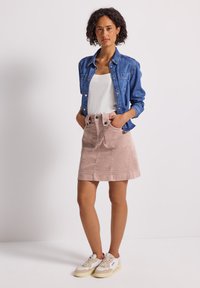 Kvinna står med händerna i fickorna, iklädd en blå denimjacka, vit topp, rosa kordkjol och vita sneakers på en enfärgad bakgrund.