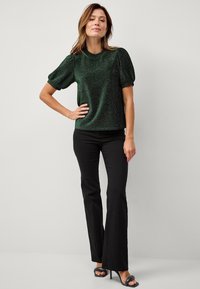 Donkergroen, sprankelend topje met korte mouwen, ronde hals en pofmouwen, gecombineerd met zwarte flared broek en zwarte haksandalen.