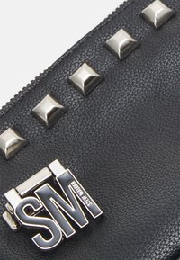 Pochette en cuir noir texturé ornée de clous argentés et d'un logo argenté et noir emblématique, présentant un design moderne avec des accents métalliques.