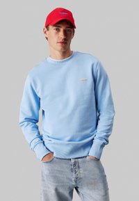 Γαλάζια φούτερ με ραμμένες μανσέτες και στρίφωμα, με μια μικρή ετικέτα στο στήθος. Συνδυασμένο με ξεθωριασμένα μπλε τζιν και κόκκινο καπέλο.