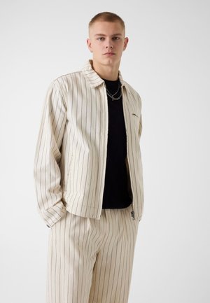 Jonge man die een crèmekleurig gestreept jasje en bijpassende broek draagt over een zwart shirt, staand met handen in de zakken tegen een effen achtergrond.