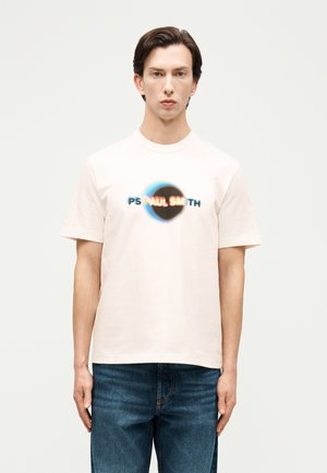 Ung mann med brunt hår iført en hvit kortermet T-skjorte med en uskarp rund "PS PAUL SMITH"-logo og blå jeans.