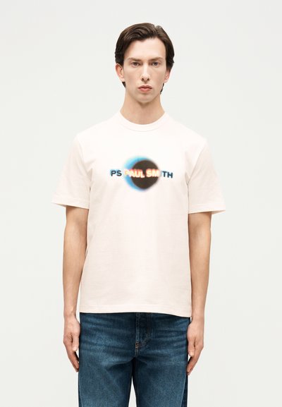 Jeune homme aux cheveux bruns portant un T-shirt blanc à manches courtes avec un logo circulaire flou "PS PAUL SMITH" et un jean bleu.