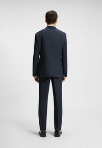 Mörkblå kostym med strukturerad tyg, slim fit-design, enkel slits bak, och matchande byxor som bärs med svarta skor.