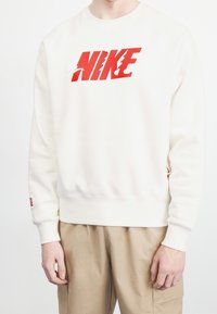Persona vestida con un sudadera blanca con un gran logo rojo "NIKE" y pantalones beige, de pie contra un fondo blanco liso.