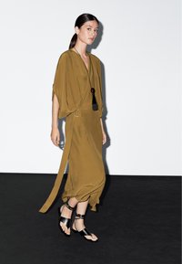 Olivově zelené, lehké šaty s křidélkovými rukávy, bočním vázáním a rozparkem. Spárovány s černými sandály na tenkých páscích. Elegantní a minimalistický design.