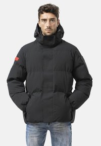 Schwarze, wattierte Jacke mit hochgestelltem Kragen, sichtbaren Nähten, einem roten Akzent am linken Ärmel und einer glatten, matten Oberfläche.