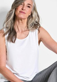 Weiße ärmellose Bluse mit rundem Kragen, glatter Textur und lockerer Passform. Ein Modell trägt sie im Sitzen und zeigt einen entspannten Stil.