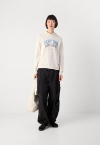 Person wearing a cream hoodie with "GANT 1949 New Haven," black cargo pants, black sneakers, holding a large cream tote bag, standing on plain white background.