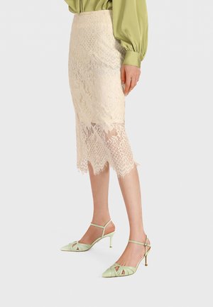 Donna che indossa una gonna midi in pizzo beige con orlo smerlato e sandali con tacco verde chiaro, in piedi contro uno sfondo neutro.