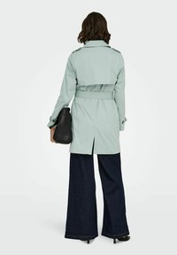 Femme aux cheveux mi-longs portant un trench-coat vert clair ceinturé, un pantalon large foncé, des talons noirs et tenant un sac à main noir.