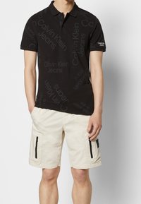 Černé polo tričko s opakujícím se logem "Calvin Klein Jeans" ve tmavě šedé barvě, spárované s béžovými cargo šortkami s dvěma zipovými kapsami.