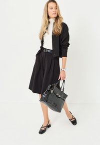Černá skládací sukně v kombinaci s bílým rolákem a černým kardiganem, doplněná lesklou černou kabelkou s texturovanými akcenty.