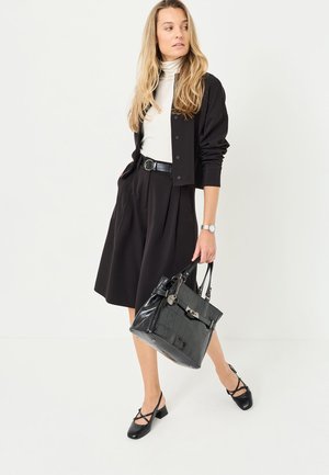 Gonna nera plissettata abbinata a un dolcevita bianco e a un cardigan nero, completata da una borsa nera lucida con dettagli testurizzati.