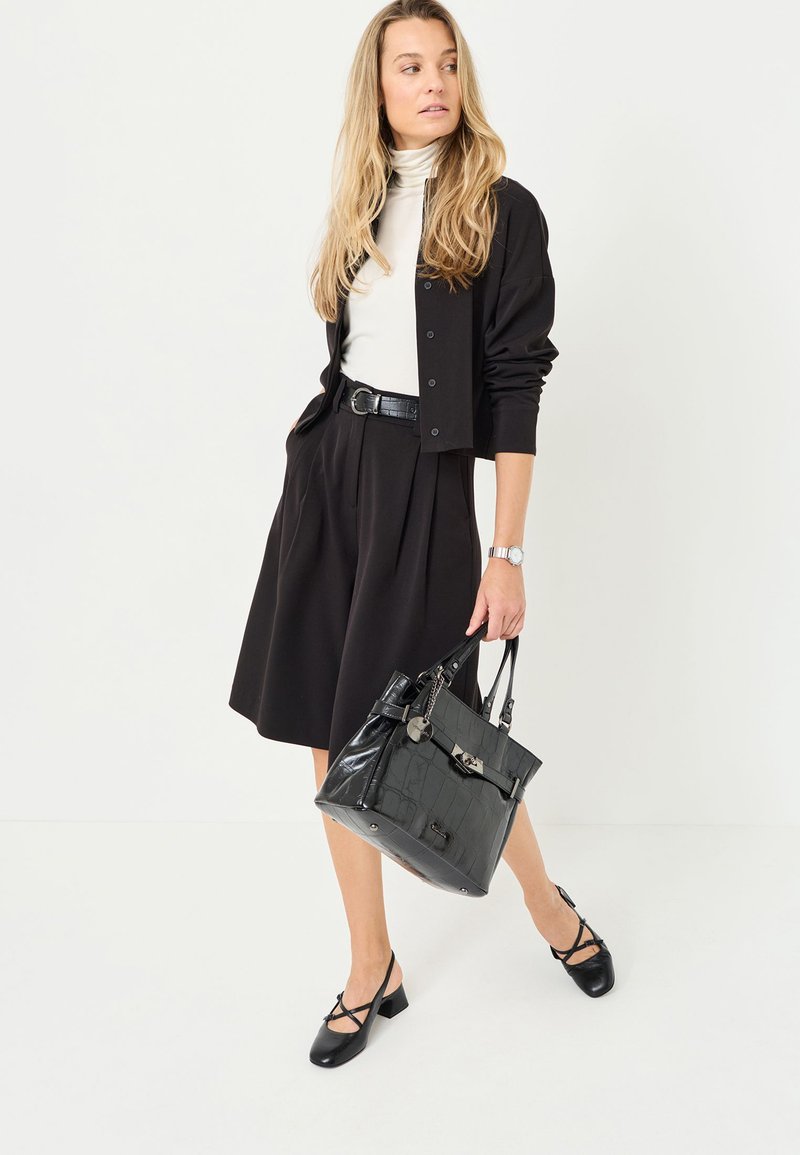 Černá skládací sukně v kombinaci s bílým rolákem a černým kardiganem, doplněná lesklou černou kabelkou s texturovanými akcenty.
