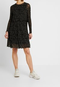 Mustapuku, jossa on pitkät hihat ja kerroksellinen muotoilu. Kuvioina valkoiset pilkut, läpikuultava kangas, ja se on yhdistetty vaaleisiin lenkkareihin.