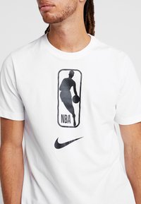 Camiseta de algodón blanca con un logo de la NBA impreso en negro y el swoosh de Nike. Diseño de mangas cortas y cuello redondo. Textura suave.