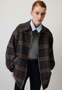 Karirasta jakna v temno modri in rjavi barvi z belimi poudarki, z zadrgo, širokim ovratnikom ter elastičnimi manšetami, oblečena čez sivi pulover.