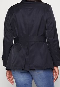 Chaqueta peplum negra con cintura ceñida, hecha de un tejido suave. Presenta una parte trasera entallada con pliegues y puños estilizados. Llevada con vaqueros azules.
