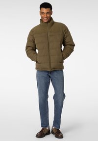 Olivengrønn puffjakke med høy krage, quiltet tekstur, glidelås foran, kombinert med blå jeans og brune sko.