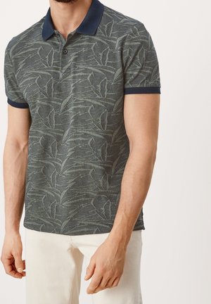 Mężczyzna ubrany w krótki rękaw, ciemnozieloną koszulkę typu polo z wzorem, granatowym kołnierzykiem oraz białe spodnie, stojący na jednolitym tle.