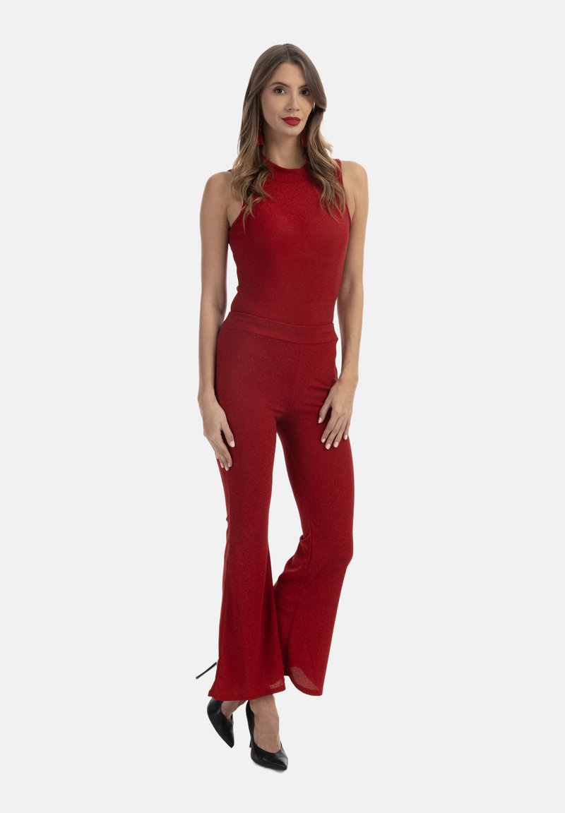 Tuta rossa senza maniche con un busto aderente e pantaloni a campana. Il tessuto ha un leggero luccichio. Tacchi alti neri completano l'outfit.