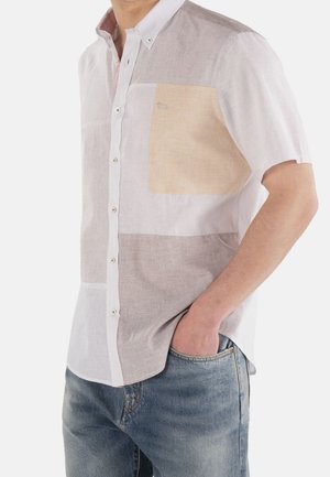 Muž nosící krátký rukáv s knoflíky a vzorem patchwork v béžové, bílé a hnědé barvě, v kombinaci s modrými džíny.