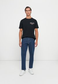 Ung mand står op, iført en sort GANT 1949 t-shirt, blå jeans og hvide sneakers mod en ensfarvet lys baggrund.