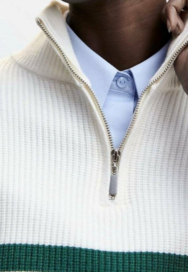 Cream-colored knit pullover with a half-zipper, featuring a green striped pattern along the hem and collar over a light blue shirt.