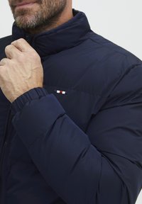 Marineblaue Pufferjacke mit hohem Kragen. Verfügt über einen Reißverschluss und elastische Bündchen. Kleines Logo in Rot, Weiß und Blau auf der Brust.