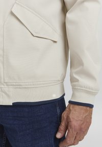 Light beige jacket with a structured design, featuring a diagonal pocket, ribbed cuffs, and a contrasting navy blue hem. Smooth texture.