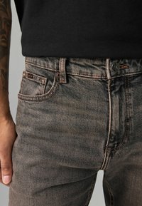 Jeans grii decolorati cu o croială slim, cusături din cupru și un singur buzunar mic cu etichetă. Închidere cu nasture și un brâu solid.