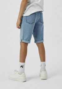 Shorts en denim bleu clair avec des ourlets retroussés, comportant des poches arrière et une coupe droite. Portés avec des baskets blanches et des chaussettes avec un logo.