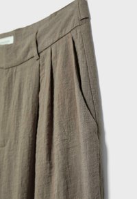 Pantalon en lin marron avec plis devant, poche latérale et taille avec passants de ceinture, présenté sur un fond clair.