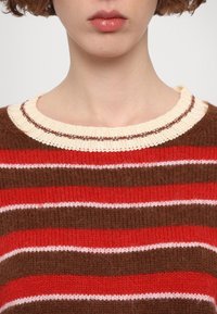 Pull en maille rayé avec un col côtelé crème et marron. Les couleurs comprennent le rouge, le marron et le blanc avec des rayures de différentes épaisseurs.