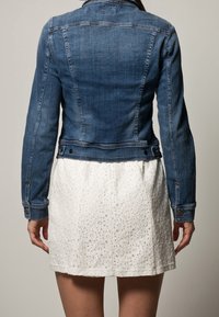 Jeansjacka med figurnära design, blå färg och knapprdetaljer, kombinerad med en vit texturerad kjol med hålmönster.