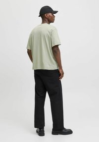 Tricou din bumbac verde deschis cu mânecă scurtă, asociat cu pantaloni negri largi și pantofi negri. Persoana poartă o șapcă neagră.