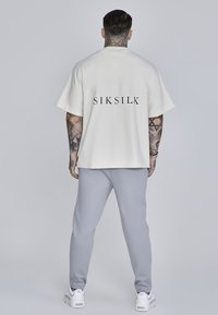 Valge ülev oversized lühikeste varrukatega T-särk musta "SIKSILK" logoga, koos heledate hallide spordipükstega ja valgete tossudega, näidates mugavat sobivust.