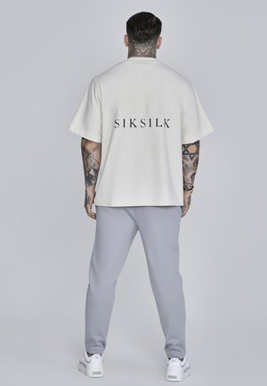 Biele prehnané tričko s krátkym rukávom s čiernym logom "SIKSILK", v kombinácii so svetlošedými teplákmi a bielymi teniskami, s uvoľneným strihom.