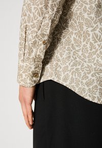 Chemise beige à manches longues avec motif de feuilles marron clair, poignets boutonnés, portée avec un pantalon noir, main posée sur le côté.