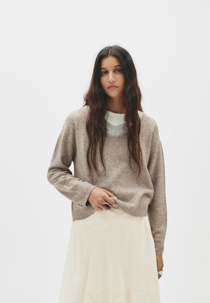 Brauner gestrickter Pullover mit V-Ausschnitt, gerippten Bündchen und Saum. Kombiniert mit einem hellen, geschichteten Top und einem fließenden cremefarbenen Rock.