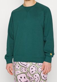 Sudadera verde con cuello redondo, puños acanalados y un acento de logo dorado en la manga. Combinada con pantalones de tonos pastel con estampados.