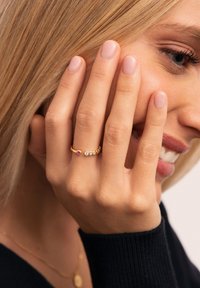 Anillo de oro con una piedra preciosa rosa y tres piedras claras dispuestas en línea, llevado en una mano con uñas naturales sobre un fondo claro.