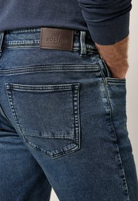 Jeans de mezclilla azul oscuro con un parche de cuero etiquetado "BOSTON PARK" en la cintura, con costuras en contraste y un diseño clásico de cinco bolsillos.