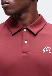 Gros plan d’une personne portant un polo bordeaux avec des boutons blancs et un "m" en cursive brodé en blanc sur la poitrine.